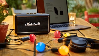 Der Marshall Bromley 450 steht draußen auf einer Dach-/Terrassenfläche neben einer Holzbank. Die Front zeigt das typische Marshall-Grillmuster mit Logo; mehrere kleine Lichtpunkte leuchten in der Front. Im Hintergrund sind Himmel, Wolken und eine Stadtansicht erkennbar.