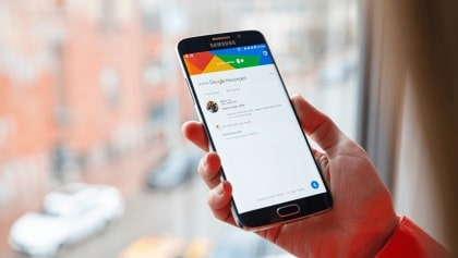 Das Bild zeigt ein modernes Smartphone, das schräg in einer Hand gehalten wird. Der Bildschirm ist eingeschaltet und zeigt eine Nachrichten-App mit dem Titel „Messages". Die Benutzeroberfläche ist überwiegend weiß gestaltet und enthält mehrere Chat-Unterhaltungen. Im oberen Bereich des Displays stehen Uhrzeit, Signal- und Akkusymbole. Darunter befindet sich die Überschrift „Messages". Anschließend folgen mehrere Nachrichtenkontakte mit kleinen Profilbildern oder Symbolen. Sichtbar sind unter anderem die Namen „Lindsey Smith", „Christina Adams" sowie ein Gruppenchat. Unter den Namen stehen kurze Vorschautexte der letzten Nachrichten. Weiter unten ist eine Nachricht von „Microsoft" zu sehen, die einen Verifizierungscode erwähnt. Rechts neben dem Smartphone schwebt ein großes blaues Symbol einer Sprechblase mit drei weißen Punkten. Das Symbol erinnert an eine Nachrichten- oder Chat-App und dient als grafische Hervorhebung des Themas Kommunikation. Der Hintergrund ist unscharf und in verschiedenen Blautönen gehalten. Mehrere große Lichtkreise erzeugen einen sogenannten Bokeh-Effekt, wodurch das Smartphone und das Chat-Symbol deutlich im Vordergrund stehen. Die Beleuchtung wirkt modern und technisch.