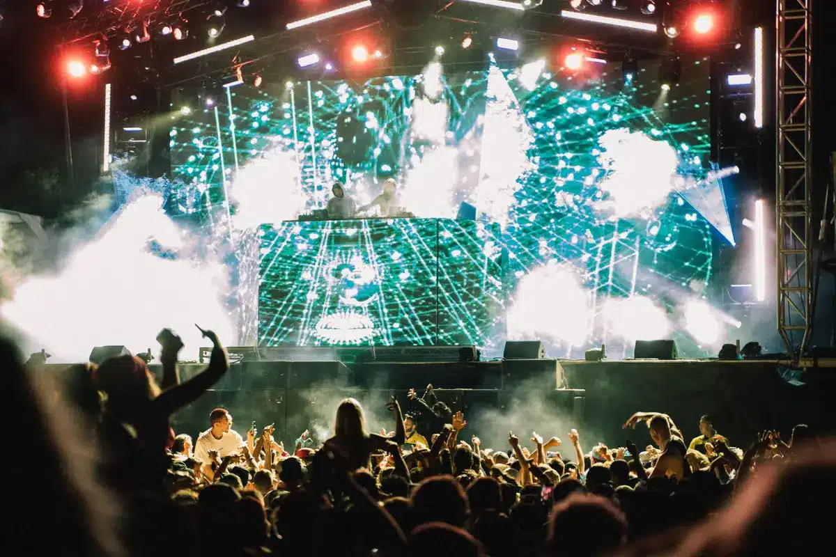 Menschenmenge mit erhobenen H&auml;nden vor B&uuml;hne mit bunten Lichtern und Pyrotechnik auf einem Musikfestival