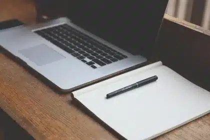 Junge Fachkraft arbeitet an Laptop mit Notizbuch und Handy auf einem Café-ähnlichen Schreibtisch